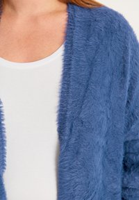 Blue fluffy cardigan made of soft, textured fabric, featuring an open front design. Worn over a plain white top.
