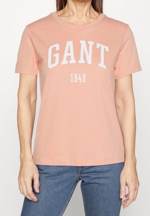 Frau trägt ein hellrosa T-Shirt der Marke GANT mit weißem Schriftzug "GANT 1949" und blaue Jeans, steht vor einem schlichten Hintergrund.