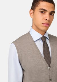 Beige pletená vesta s výstrihom do V, s gombíkmi a hladkou textúrou, nosená cez svetlomodrú košeľu s golierom a vzorovanú hnedú kravatu.