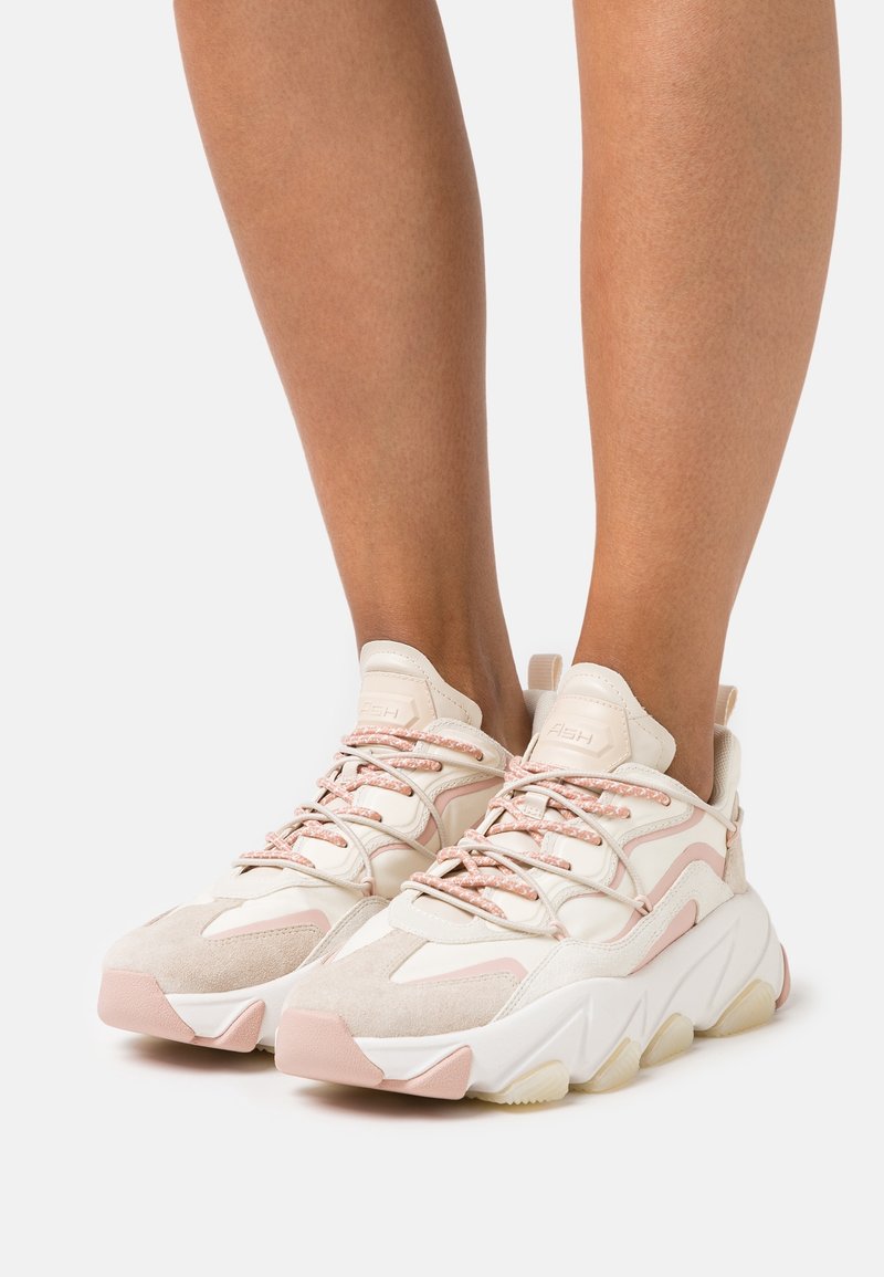 Jambes portant des baskets blanches et rose pâle épaisses avec des semelles épaisses et des lacets à motifs sur un fond neutre.