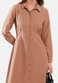 Vestito camicia di un caldo colore marrone, con maniche lunghe, colletto e un tessuto liscio e testurizzato. Dettagli di cucitura sottili.