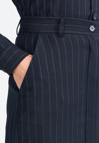 Tessuto a righe blu navy con cuciture chiare in contrasto, caratterizzato da una vita aderente e tasche. Il materiale appare liscio e strutturato.
