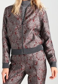 Bordeaux bomberjack met metallic bloemmotief, grijze ribgebreide boorden en zoom, met ritssluiting en lichte, gladde textuur.