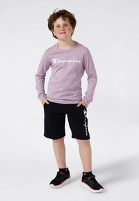 Világos lila hosszú ujjú ing fehér "Champion" logóval, fekete rövidnadrággal, amelyen függőleges logóminta található, és fekete sportcipővel párosítva.