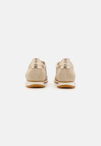 Beige mokasiner i nubuck med metalliske detaljer, rund tå og hvit gummisåle. Har en teksturert bakdel og kontrastfargede snører.