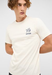 Cream-colored cotton t-shirt with short sleeves, featuring a blue graphic of a horse and the word "MUSTANG" on the left chest area.