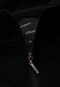 Chaqueta negra con cremallera hecha de tejido suave, con una cremallera de metal y un tirador de marca, además de una etiqueta interior con la talla M.