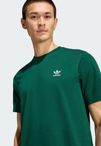 Zelené bavlnené tričko s okrúhlym výstrihom, krátkymi rukávmi a malým bielym logom Adidas na hrudi. Hladká textúra.