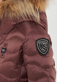 Chaqueta de plumas marrón con diseño acolchado, que cuenta con una capucha forrada de piel y una insignia negra con lentejuelas en el hombro.
