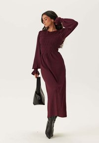 Robe longue bordeaux à manches longues avec un corsage smocké et une jupe fluide. Un sac à main noir et des bottes jusqu'aux genoux complètent l'ensemble. Tissu texturé.