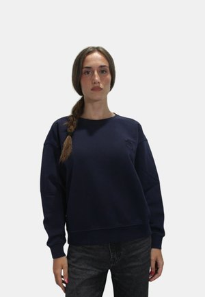 Felpa blu navy con collo a giro e polsini a costine, con logo ricamato sul petto. Abbinata a jeans scuri.