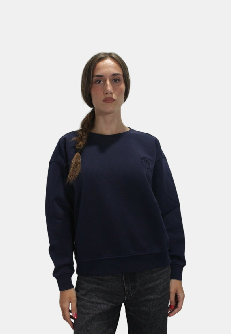 Felpa blu navy con collo a giro e polsini a costine, con logo ricamato sul petto. Abbinata a jeans scuri.
