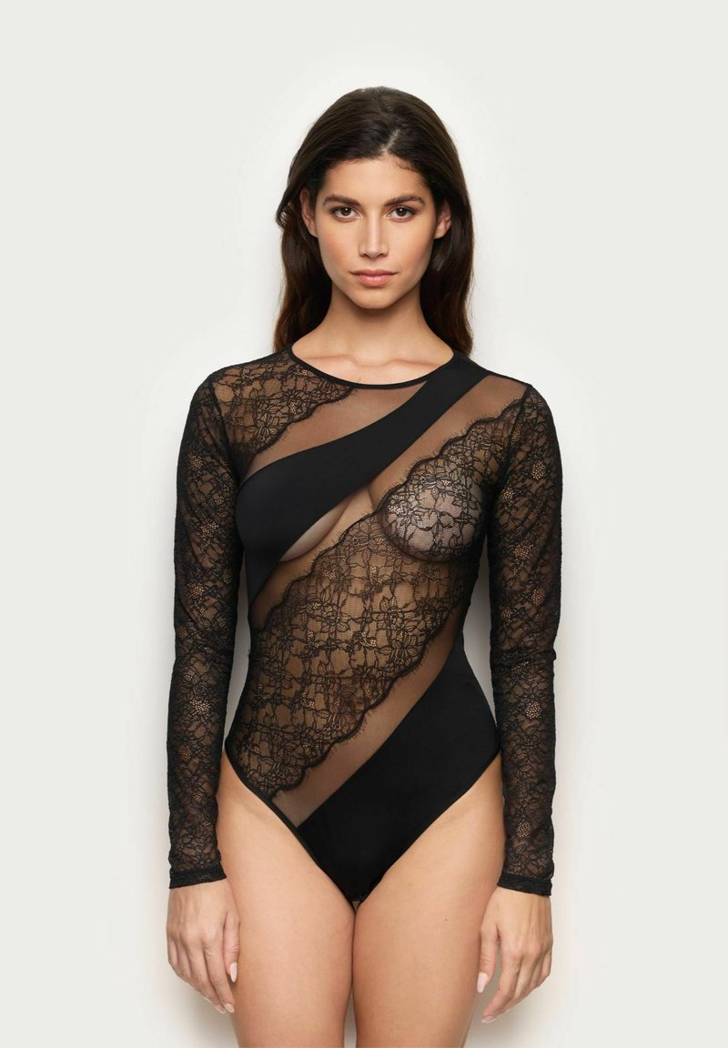 Žena v černém asymetrickém body s průhlednými krajkovými panely a dlouhými rukávy, stojící na pozadí v jemných svetel.