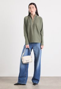 Eine Person steht vor einem neutralen Hintergrund, trägt einen grünen Reißverschluss-Top und weit geschnittene Jeans und hält eine kleine beigefarbene Handtasche.