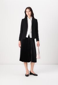 Mladá žena stojí proti bílému pozadí, obléká černý blazer, bílou košili a plisovanou černou sukni, drží malou růžovou kabelku.