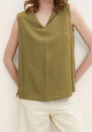 Vrouw draagt een mouwloze olijfgroene blouse met kraag, gecombineerd met gebroken witte broek, armen op de heupen.