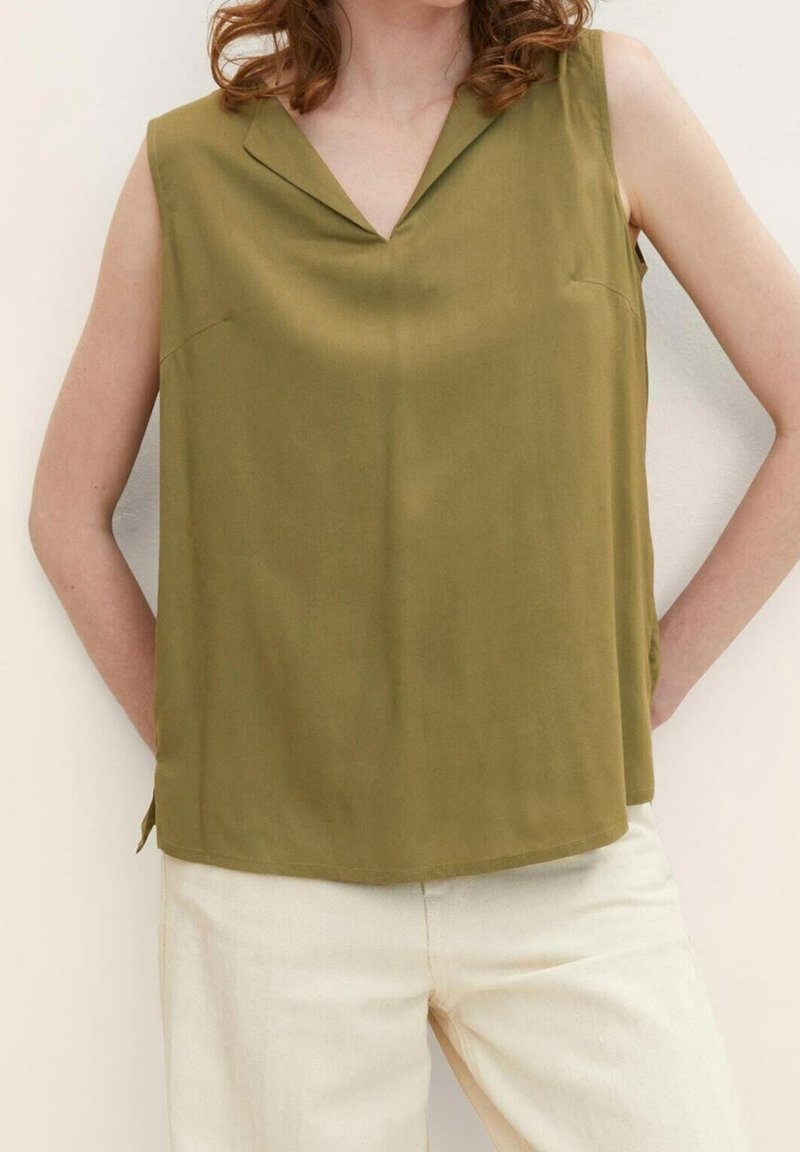 Femme portant un chemisier sans manches vert olive avec col, assorti à un pantalon blanc cassé, les bras posés sur les hanches.