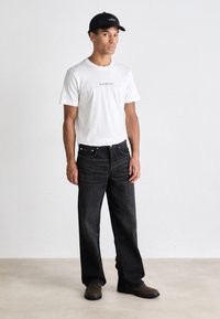 Weißes Baumwoll-T-Shirt mit schwarzem Logo, kombiniert mit locker sitzenden schwarzen Jeans und braunen Schuhen. Das Model trägt eine schwarze Mütze.