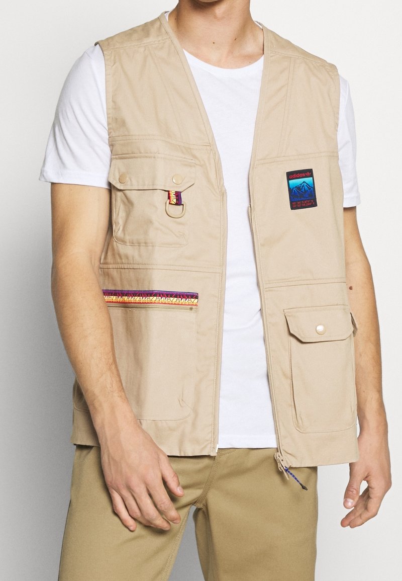 Beige lättviktsväst med flera fickor, med en färgglad dekorerande remsa och logotyp-patch, bärs över en vit T-shirt.