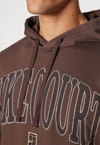 Sweatshirt com capuz castanho e grande texto branco "KICKCOURT". Apresenta um tecido texturizado, capuz com cordão e detalhe do logótipo da Nike.