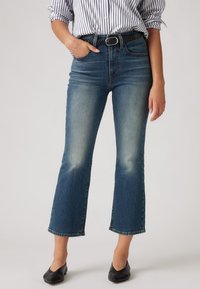 Donna che indossa jeans flare di media altezza blu con una cintura nera intrecciata, una camicia a righe con maniche arrotolate e ballerine nere a punta.