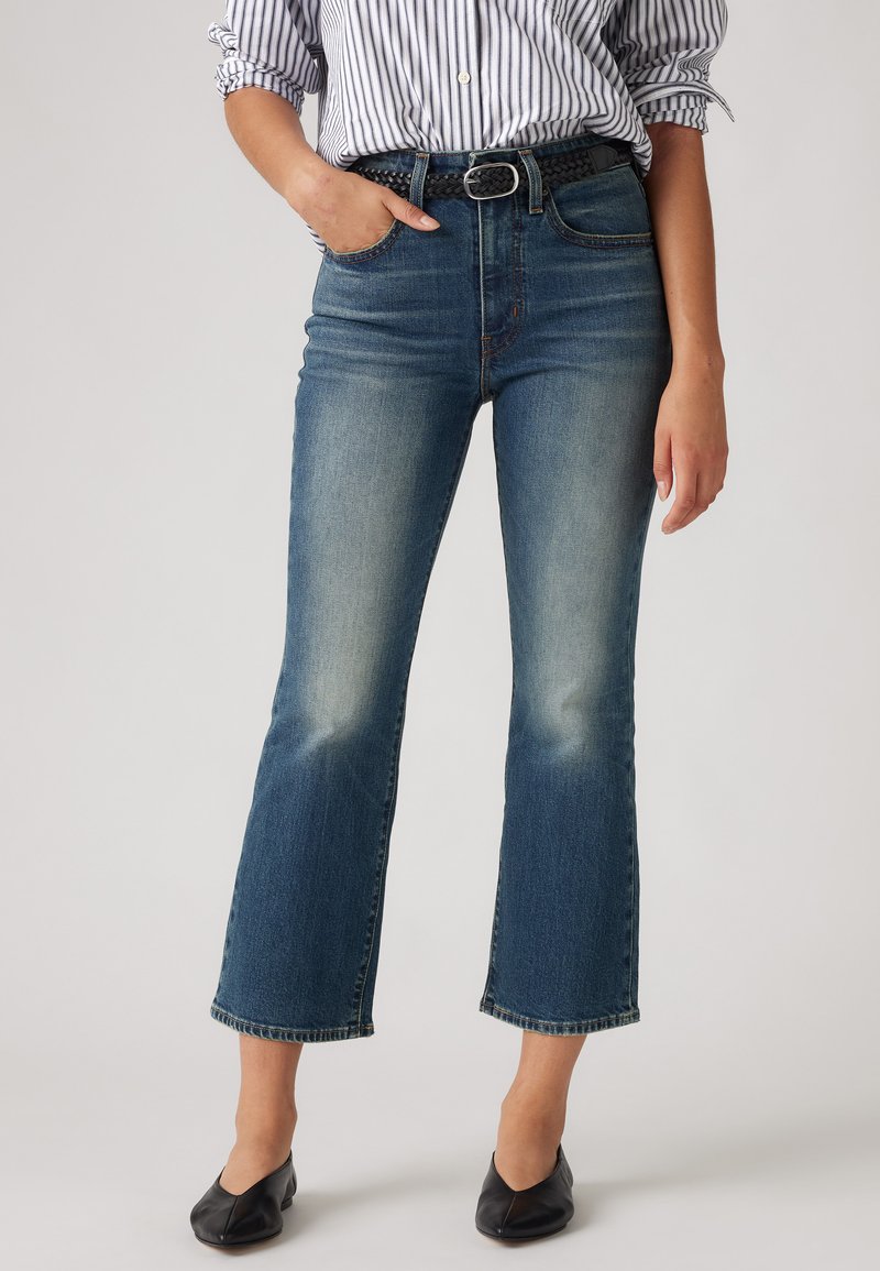 Donna che indossa jeans flare di media altezza blu con una cintura nera intrecciata, una camicia a righe con maniche arrotolate e ballerine nere a punta.