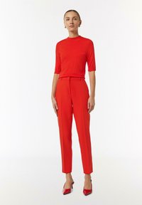 Red short-sleeve top paired with high-waisted red trousers. Both have a smooth texture. Model wears glossy red heels. Minimalist background.