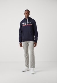 Jūras spēku kapuce ar baltu un sarkanu Champion logo, priekšējo kabatu un auklu. Saskaņota ar gaiši pelēkiem sporta biksēm un baltām sporta kurpēm.