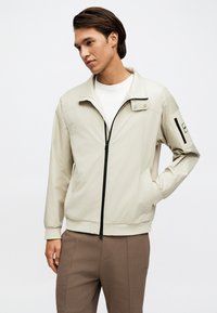 Άνδρας με μπεζ ζακέτα με φερμουάρ και ψηλό γιακά, και καφέ παντελόνι, όρθιος με το ένα χέρι στην τσέπη, σε μονόχρωμο φόντο.