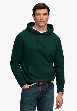 Mand iført mørkegrøn hættetrøje, blå jeans, brunt bælte og armbåndsur, stående med den ene hånd i lommen mod hvid baggrund.
