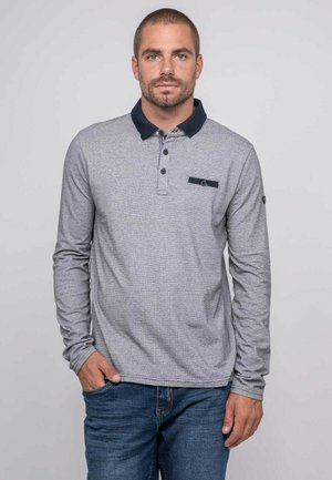 Homme aux cheveux courts et barbe portant un polo gris à manches longues avec un col foncé, debout avec une main dans la poche de son jean, sur un fond uni.