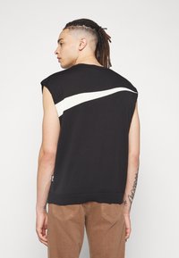 Schwarzes ärmelloses Stricktop mit einem weißen diagonalen Streifen über den Rücken. Enthält ein Logoetikett am Saum. Glatte Textur.