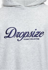 Sweat à capuche gris clair avec texte brodé bleu marine "Dropsize" en cursive et "STUDIO | COLLECTION" en majuscules en dessous.