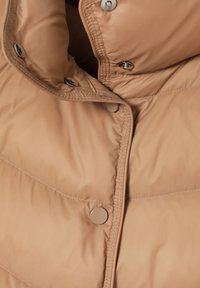 Pufferjack in zachte beige stof, met een hoge kraag, zichtbare stiksels en metalen drukknoopjes aan de voorkant.