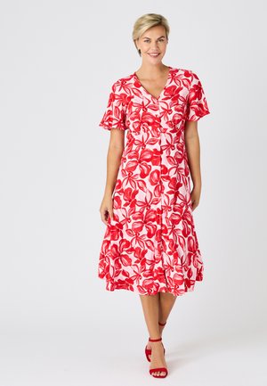 Mujer con un vestido midi blanco con estampado floral rojo, mangas cortas acampanadas, escote en V y sandalias rojas de tacón alto, de pie y sonriendo.