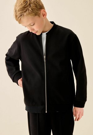 Dreng med kort blond hår iført en sort lynlåstrøje over en hvid skjorte, kigger nedad med den ene hånd på hoften mod en ensfarvet baggrund.
