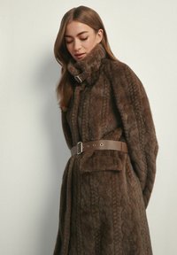 Brązowy płaszcz z futrzanym sztucznym futrem o teksturze warkocza, z wysokim kołnierzem i paskiem w talii. Posiada prostokątną kieszeń oraz srebrzysty element z klamrą.