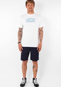 Camiseta de algodón blanca con el logo azul de "Oxbow", combinada con pantalones cortos azul marino. El modelo lleva zapatillas negras con detalles blancos.