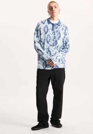 Langærmet pullover i lyseblå med et tekstureret slangeskinds mønster. Parret med sorte løstsiddende bukser og sorte sko.