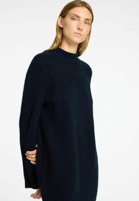 Tumši jūras zilas krāsas pārāk liels svārsts ar augstu apkakli, pazeminātiem pleciem un mīkstu, pūkainu tekstūru. Iekļauj garas piedurknes un brīvu siluetu.