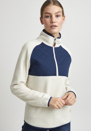 Hvit fleece genser med et marineblå quiltet panel og kvart glidelås-design, med marineblå detaljer og en liten logo nær sømmen.