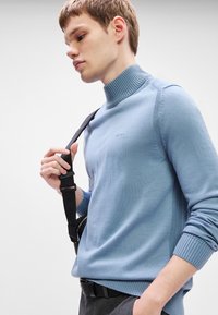 Νεαρός άνδρας φορώντας ελαφριά μπλε ζιβάγκο με το λογότυπο "BOSS", κρατώντας μια μαύρη τσάντα ώμου στον ώμο του, μπροστά σε απλό φόντο.