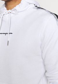 Nahaufnahme einer Person, die einen weißen Kapuzenpullover mit Kordelzügen trägt, der ein schwarzes "Champion"-Logo auf der Brust und ein markiertes Band entlang der Schulter aufweist.