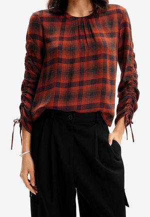 Vrouw draagt een roestkleurige en zwarte geruite blouse met geplooide mouwen, gecombineerd met een zwarte broek met hoge taille, hand in de zak.