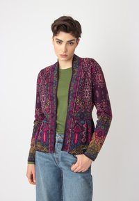 Gilet multicolore avec des motifs floraux et géométriques complexes dans des nuances de violet, rose et vert, avec une coupe droite et des poches.