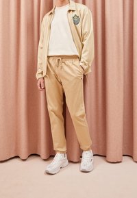 Béžová pruhovaná košile s logem, krémové tričko, béžové zúžené kalhoty a bílé tenisky. Měkká textilie, uvoln ěný střih a elastické manžety.