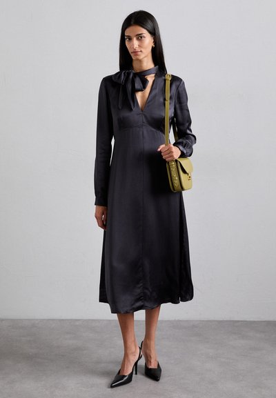 Langes, dunkles Satin-Kleid mit V-Ausschnitt und Schleifchen-Detail am Kragen. Kombiniert mit einer grünen Schultertasche und schwarzen spitzen Pumps.
