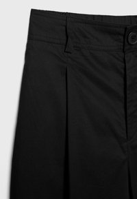 Pantalon noir taille haute avec fermeture par bouton, passants de ceinture et détail pli devant sur fond clair uni.