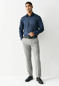 Mężczyzna w ciemnoniebieskiej koszuli zapinanej na guziki, jasnoszarych spodniach, czarnym pasku i czarnych butach eleganckich stoi na tle jednolitego białego tła.