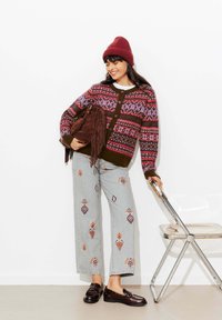 Gilet en tricot aux motifs bordeaux et violets, associé à un jean gris clair brodé. Sac à franges en daim marron et mocassins noirs.